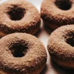 Four baked apple cider donuts coated in cinnamon sugar on a white plate, soft and golden brown, perfect for cozy fall baking.