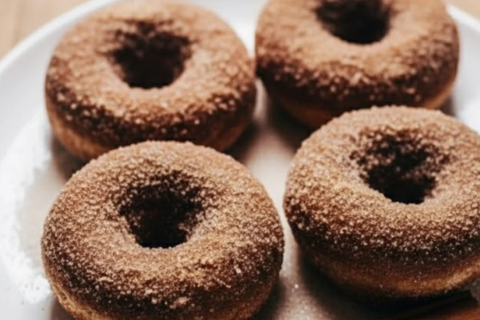 Four baked apple cider donuts coated in cinnamon sugar on a white plate, soft and golden brown, perfect for cozy fall baking.