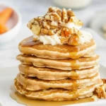 Stack of fluffy carrot cake pancakes topped with whipped cream, chopped walnuts, and maple syrup on a white plate.