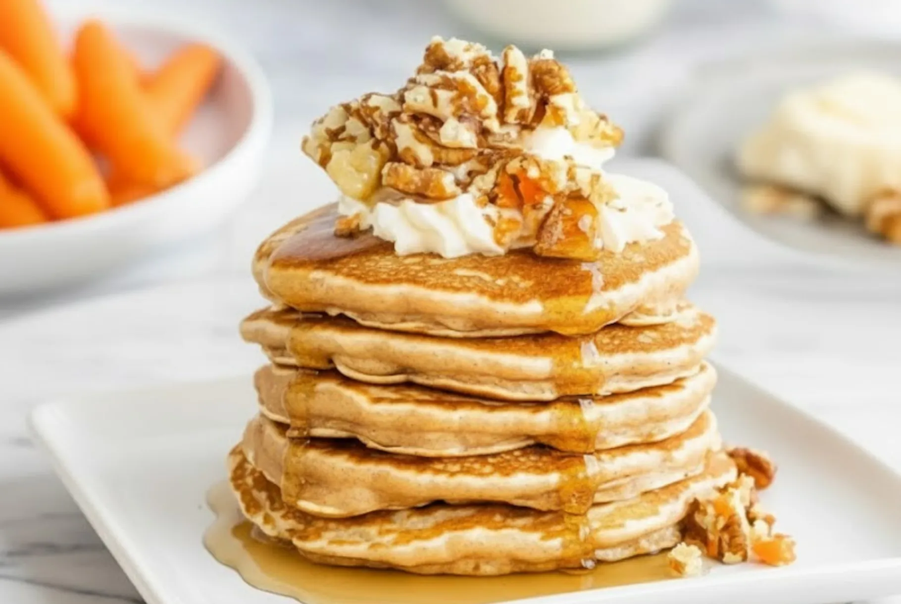 Stack of fluffy carrot cake pancakes topped with whipped cream, chopped walnuts, and maple syrup on a white plate.