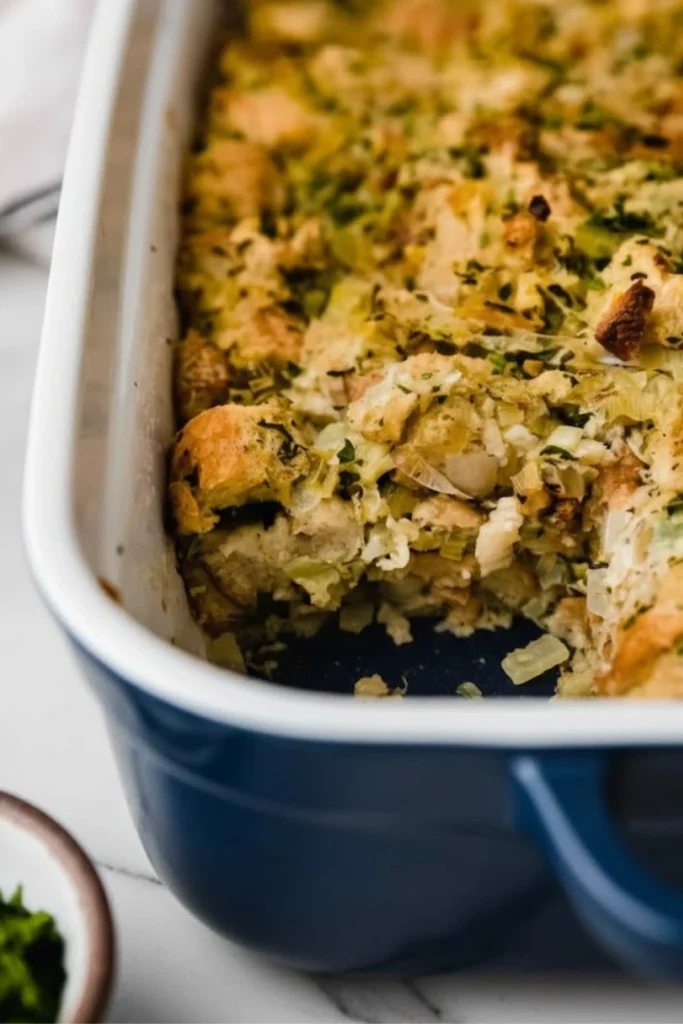 Close-up of baked herb stuffing for Thanksgiving in a blue casserole dish, featuring golden bread cubes, leeks, and fresh parsley.