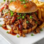 Slow Cooker Honey Garlic Pulled Pork sandwich with barbecue sauce, topped with green onions, served with fries and coleslaw on a white plate