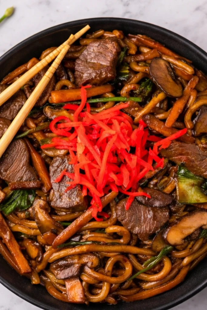 Overhead shot of beef yaki udon with stir-fried vegetables, tender beef slices, and red pickled ginger in a black bowl with chopsticks