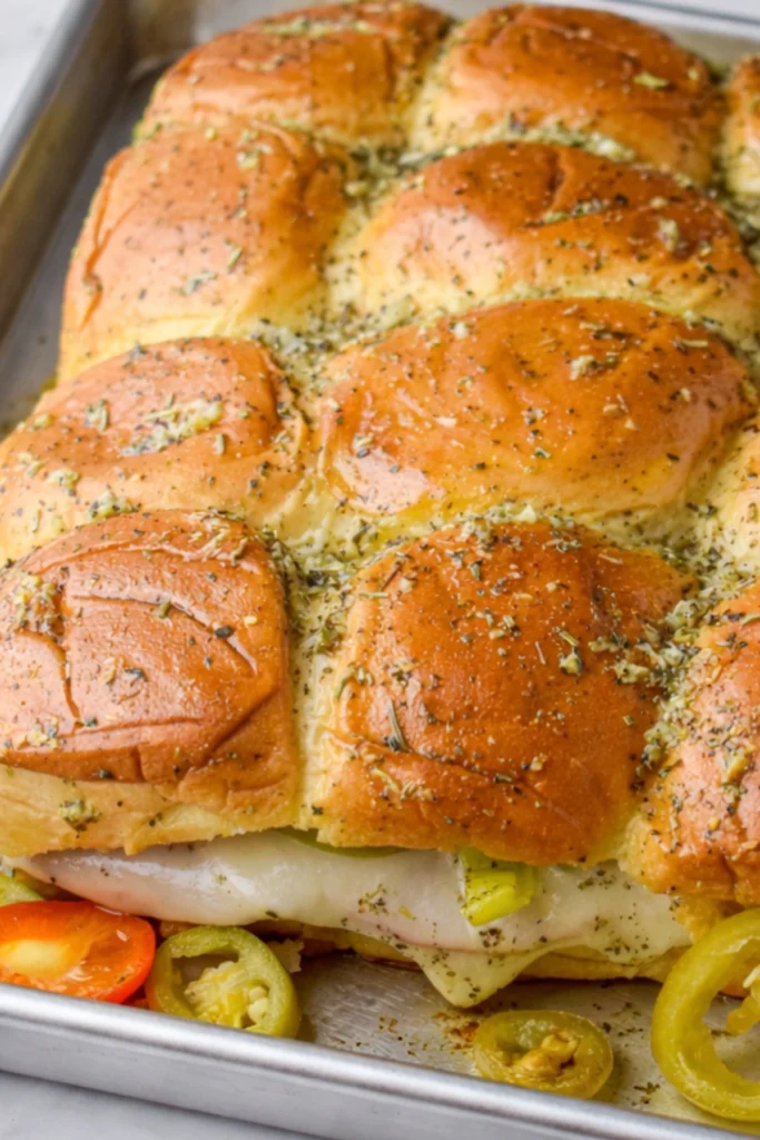 Hot Italian sub sliders baked in a pan with garlic butter rolls, melted provolone, deli meats, and sliced peppers