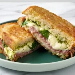 Grilled sourdough sandwich with prosciutto, mozzarella, fresh basil, and pesto on a green plate