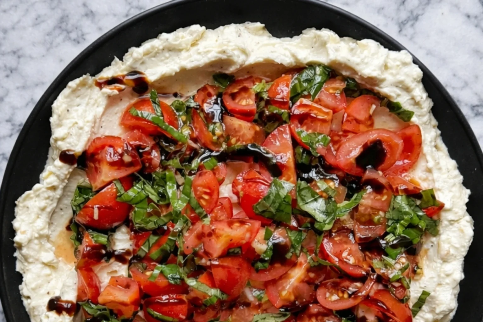 Whipped feta bruschetta dip topped with cherry tomatoes basil and balsamic glaze on a black plate