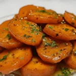 Honey glazed carrot coins stacked on a glass plate, glistening with a rich butter-honey glaze and sprinkled with fresh parsley for a vibrant, glossy finish.