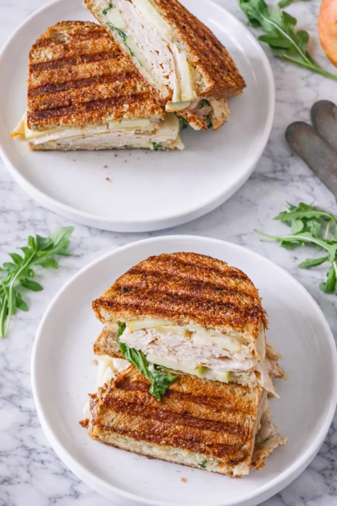 Turkey apple brie panini cut in half with melted brie cheese sliced turkey crisp apples and arugula on toasted golden bread