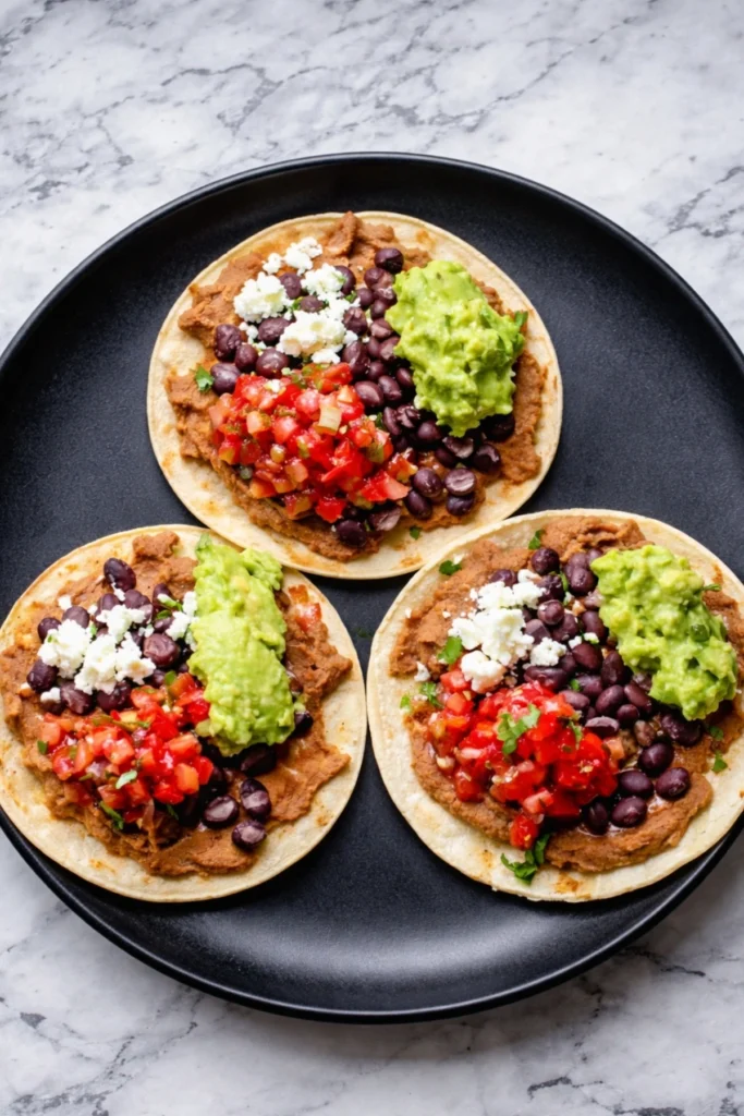 Three vegetarian tostadas with refried beans, black beans, guacamole, pico de gallo, and crumbled cheese served on a black plate