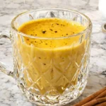 Golden Milk in a clear glass mug topped with black pepper, with cinnamon sticks on a marble counter