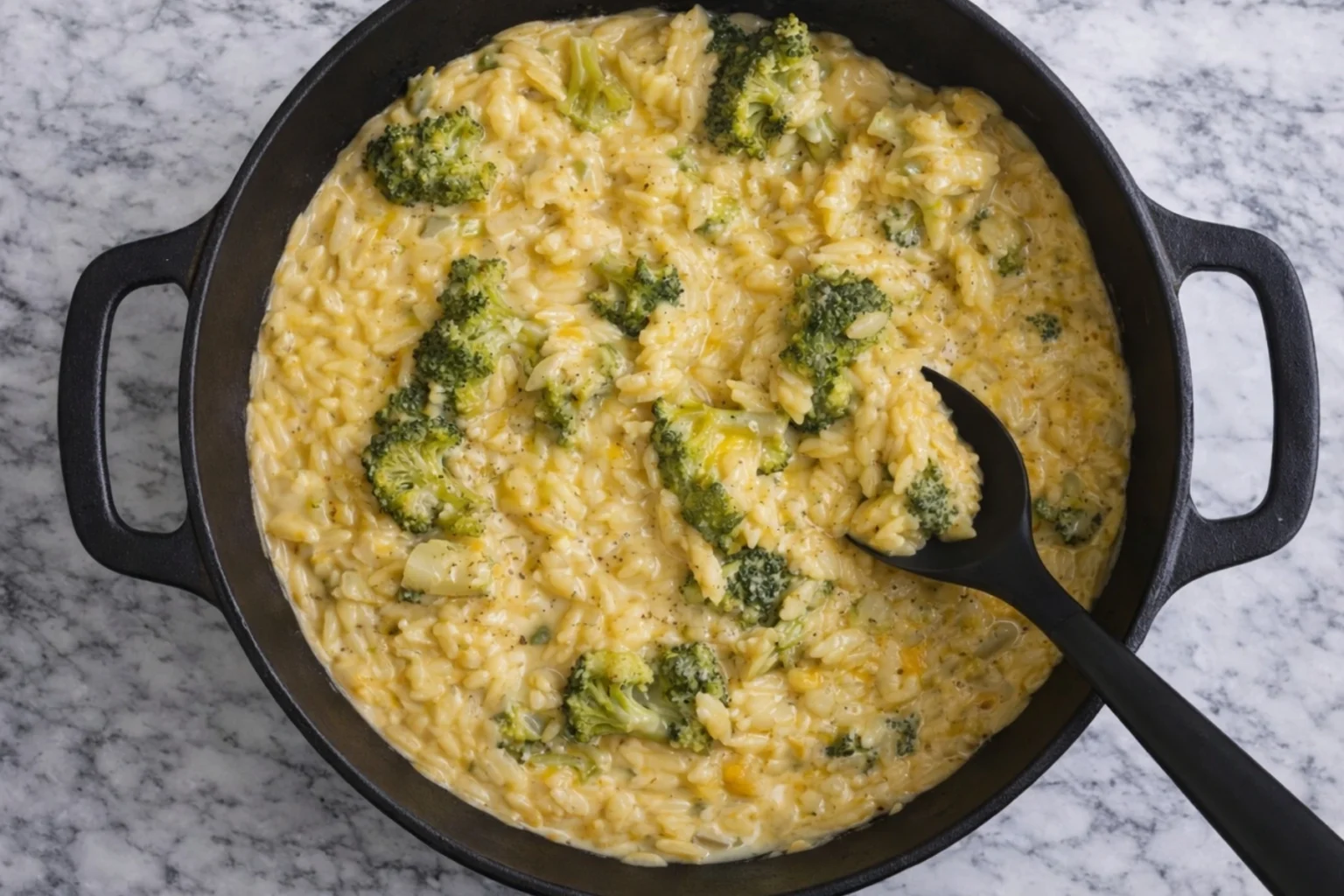 Creamy broccoli cheddar orzo in a black pot with a serving spoon on a marble countertop