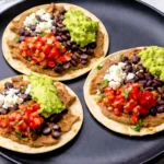 Vegetarian tostadas with refried beans, black beans, guacamole, pico de gallo, and crumbled cheese on a black plate