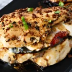Caprese stuffed chicken breast filled with melted mozzarella, sun-dried tomatoes, and basil, topped with garlic and balsamic glaze on a black plate