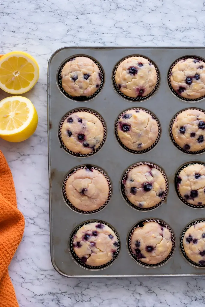 Sourdough discard blueberry lemon muffins in muffin tin with fresh lemons on marble counter