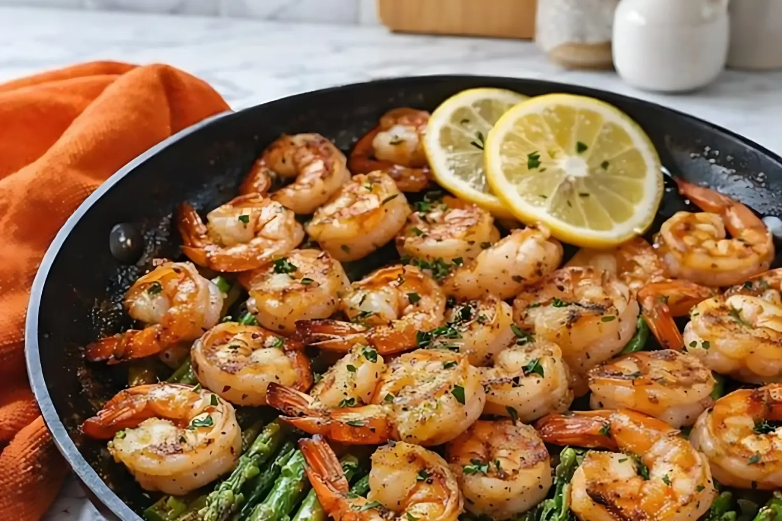Lemon garlic shrimp and asparagus in a black skillet topped with fresh lemon slices and parsley on a marble countertop