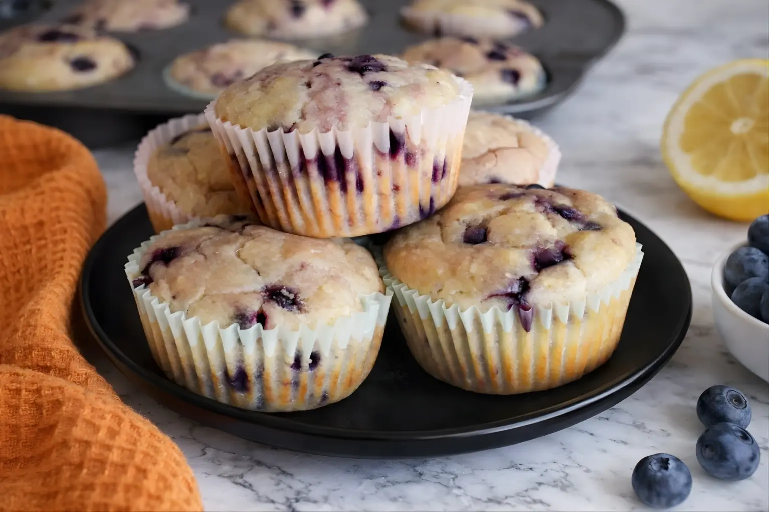 Sourdough discard blueberry lemon muffins stacked on black plate with fresh blueberries and lemon