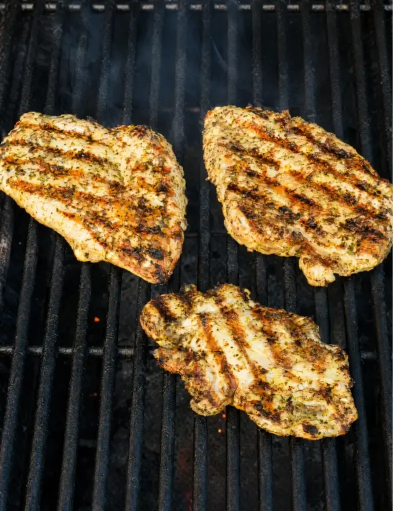 Marinated chicken breasts grilling on hot black grill grates with golden char marks, dried herbs, and light smoke for grilled chicken recipes.