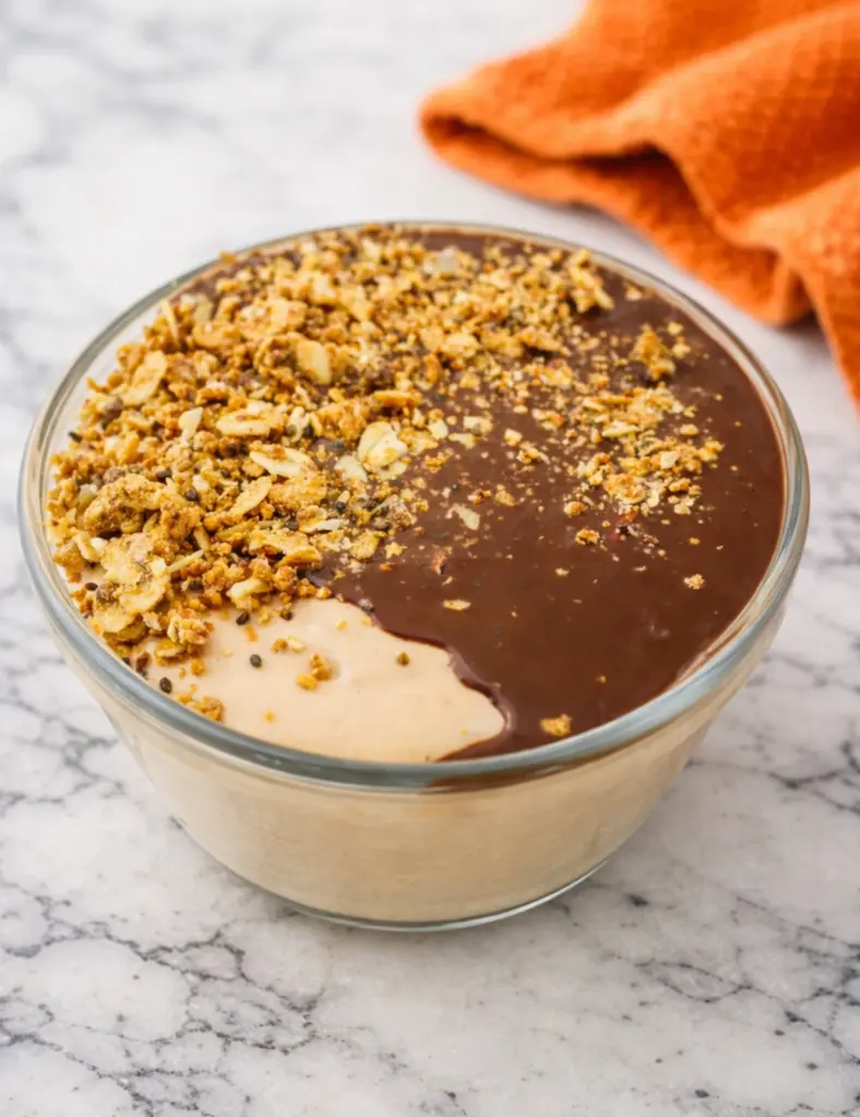 Chocolate peanut butter cottage cheese breakfast bowl topped with granola and chocolate sauce in a glass bowl on a marble countertop