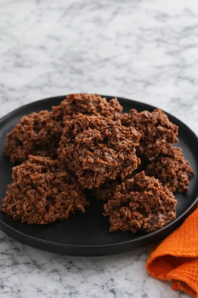 Chocolate haystack cookies stacked on black plate with orange towel on marble surface