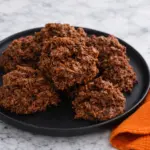 Chocolate haystack cookies on black plate with orange towel on marble countertop