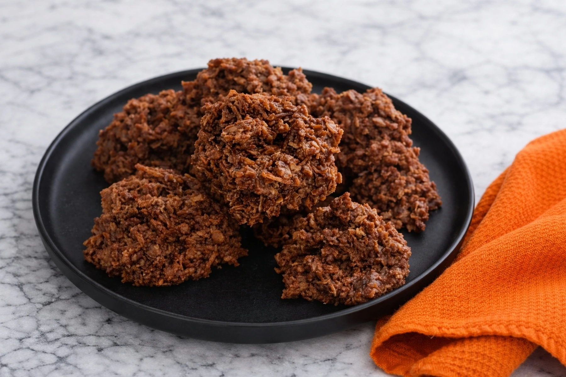 Chocolate haystack cookies on black plate with orange towel on marble countertop