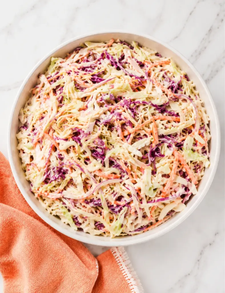 Overhead view of Creamy Coleslaw Recipe in a white bowl with shredded cabbage, carrots, red onion, and creamy mayonnaise dressing on a marble countertop.