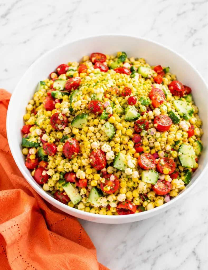 Corn salad recipe in a white bowl with fresh corn, cherry tomatoes, cucumber, bell peppers, herbs, and crumbled cheese on a marble countertop with an orange towel.