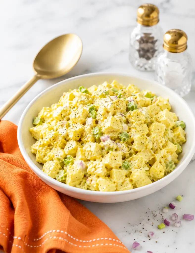 Classic potato salad in a white serving bowl with creamy mustard dressing, celery, red onion, black pepper, gold spoon, crystal shakers, and orange towel.