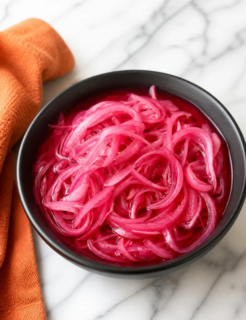 Bowl of bright pink pickled red onions on a white marble countertop with an orange kitchen towel nearby.