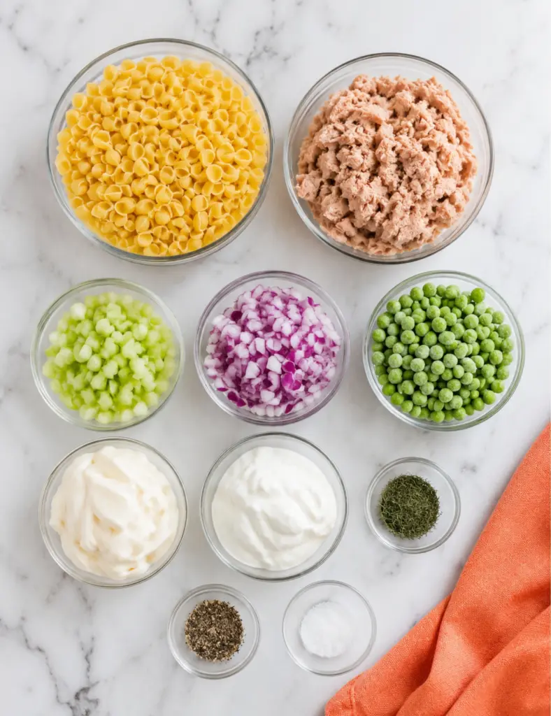 Tuna pasta salad ingredients in glass bowls with shell pasta, canned tuna, celery, red onion, peas, mayonnaise, Greek yogurt, dill, salt, and pepper on a marble countertop.