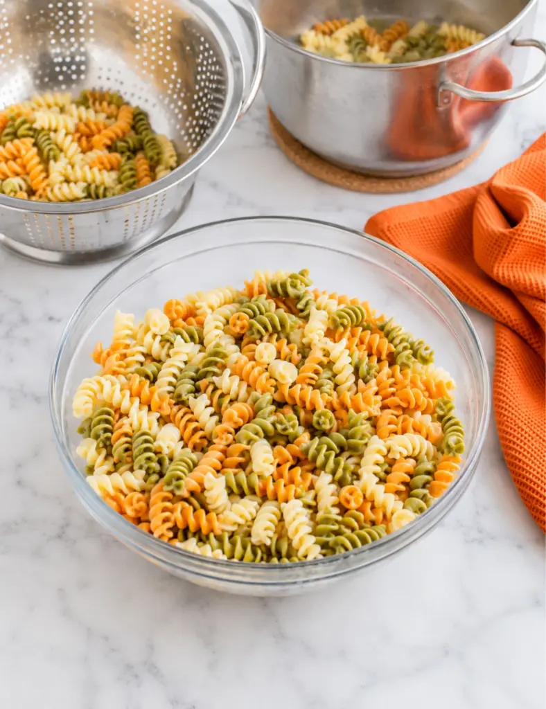Cooked tri-color rotini pasta cooling in a glass bowl with a colander, stainless steel pot, and orange towel on a white marble countertop