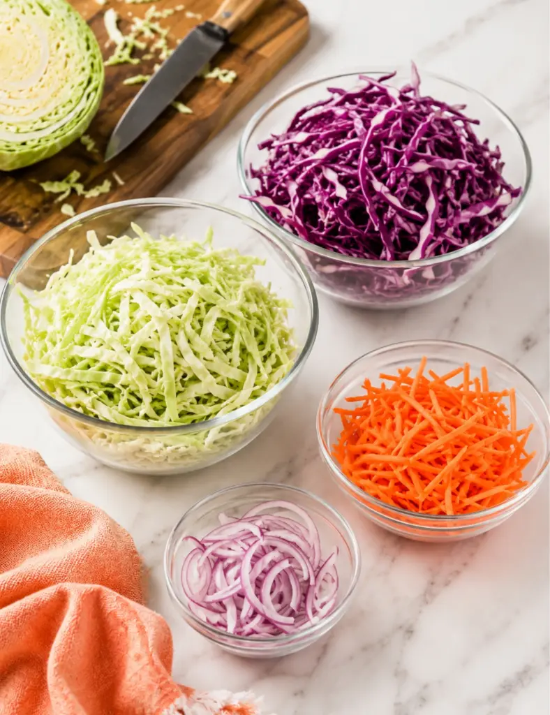 Creamy Coleslaw Recipe prep with shredded green cabbage, red cabbage, carrots, and sliced red onion in glass bowls on a marble countertop.