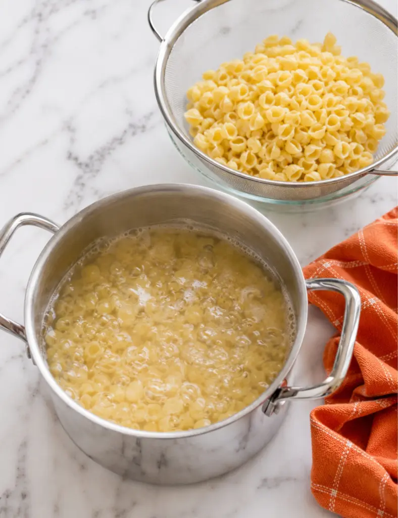 Small shell pasta boiling in a stainless steel pot with cooked pasta draining in a mesh strainer on a white marble countertop.