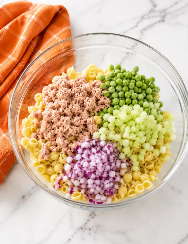 Tuna pasta salad ingredients in a glass mixing bowl with shell pasta, canned tuna, peas, celery, and red onion on a white marble countertop.