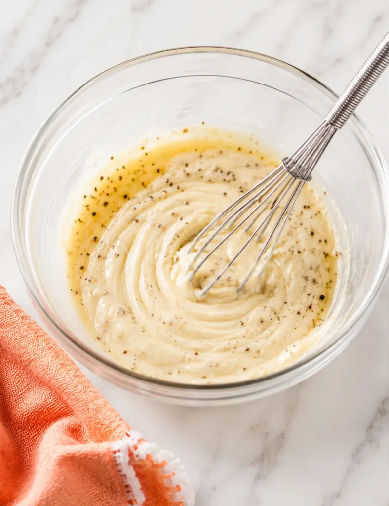 Creamy Coleslaw Recipe dressing being whisked in a glass bowl with mayonnaise, apple cider vinegar, Dijon mustard, sugar, celery seeds, salt, and black pepper.