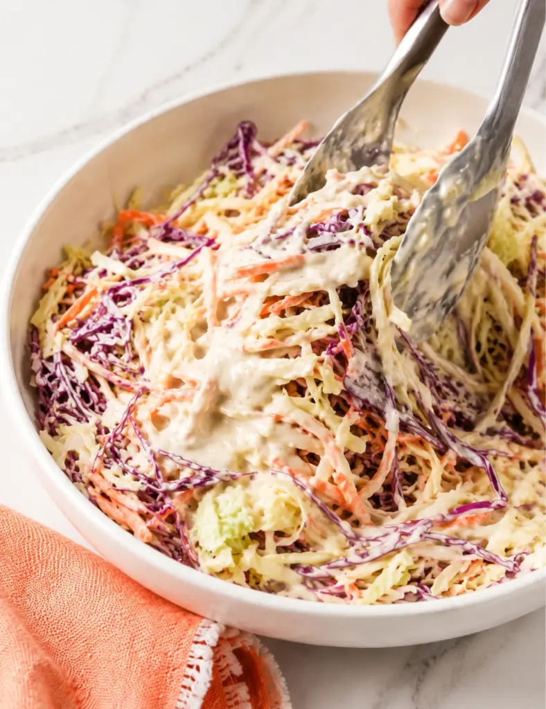 Creamy Coleslaw Recipe being tossed with tongs in a white bowl, showing shredded cabbage, carrots, and red onion coated in a creamy mayonnaise dressing.