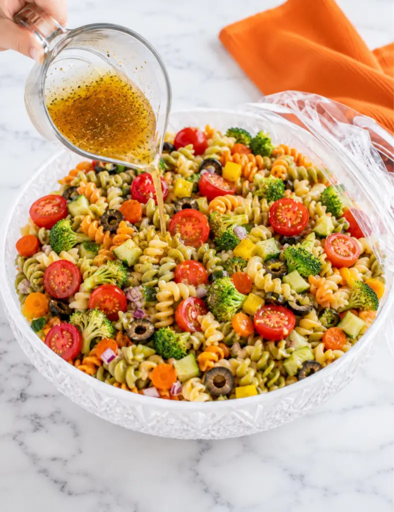 Italian dressing being poured over Pasta Salad with Veggies in a crystal bowl with rotini, tomatoes, broccoli, olives, cucumber, carrots, and orange towel