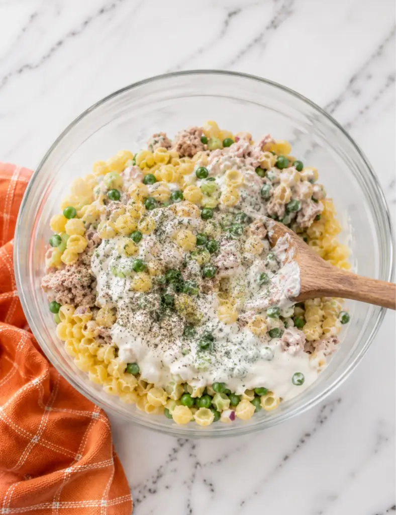 Tuna pasta salad being mixed with creamy dressing, dill, black pepper, shell pasta, tuna, peas, celery, and red onion in a glass bowl.
