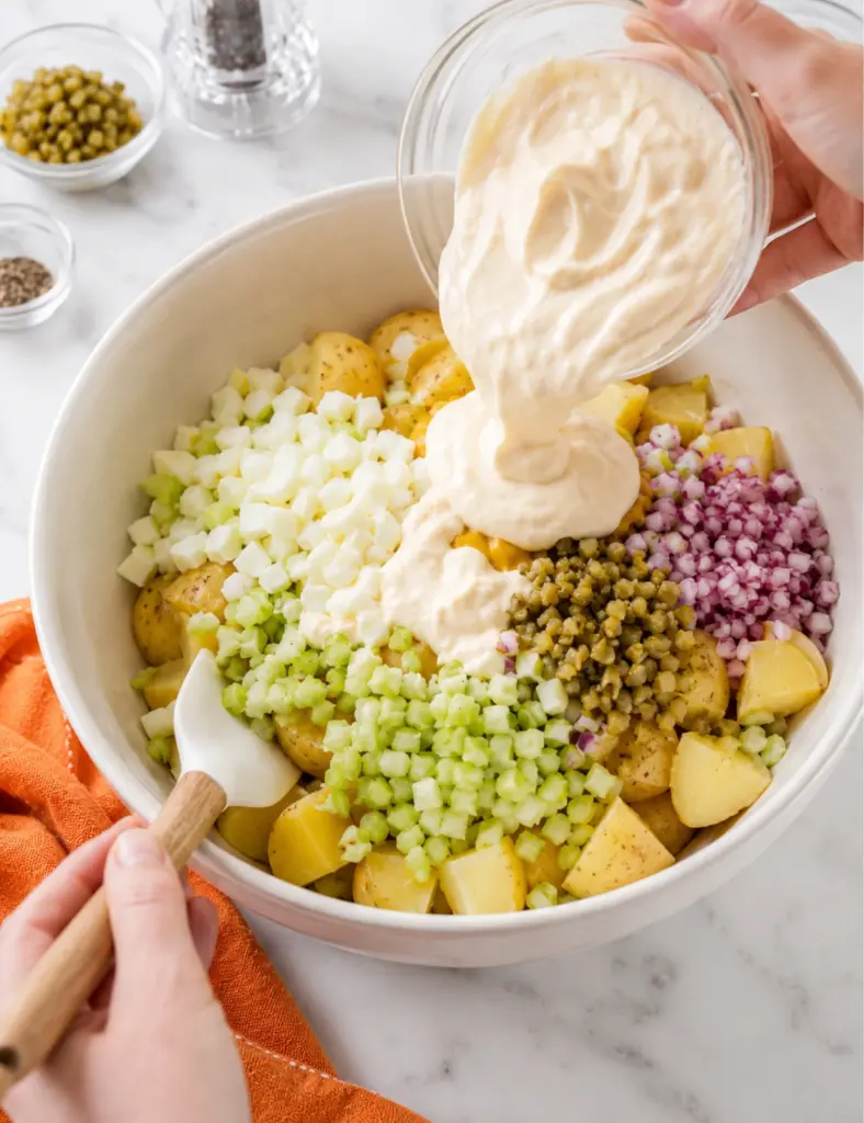 Classic potato salad being assembled in a large bowl with Yukon Gold potatoes, chopped egg whites, celery, red onion, pickles, and creamy dressing poured on top.