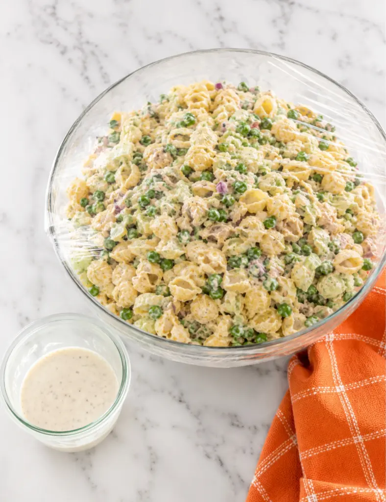 Creamy tuna pasta salad covered with plastic wrap in a glass bowl with extra dressing on a white marble countertop.