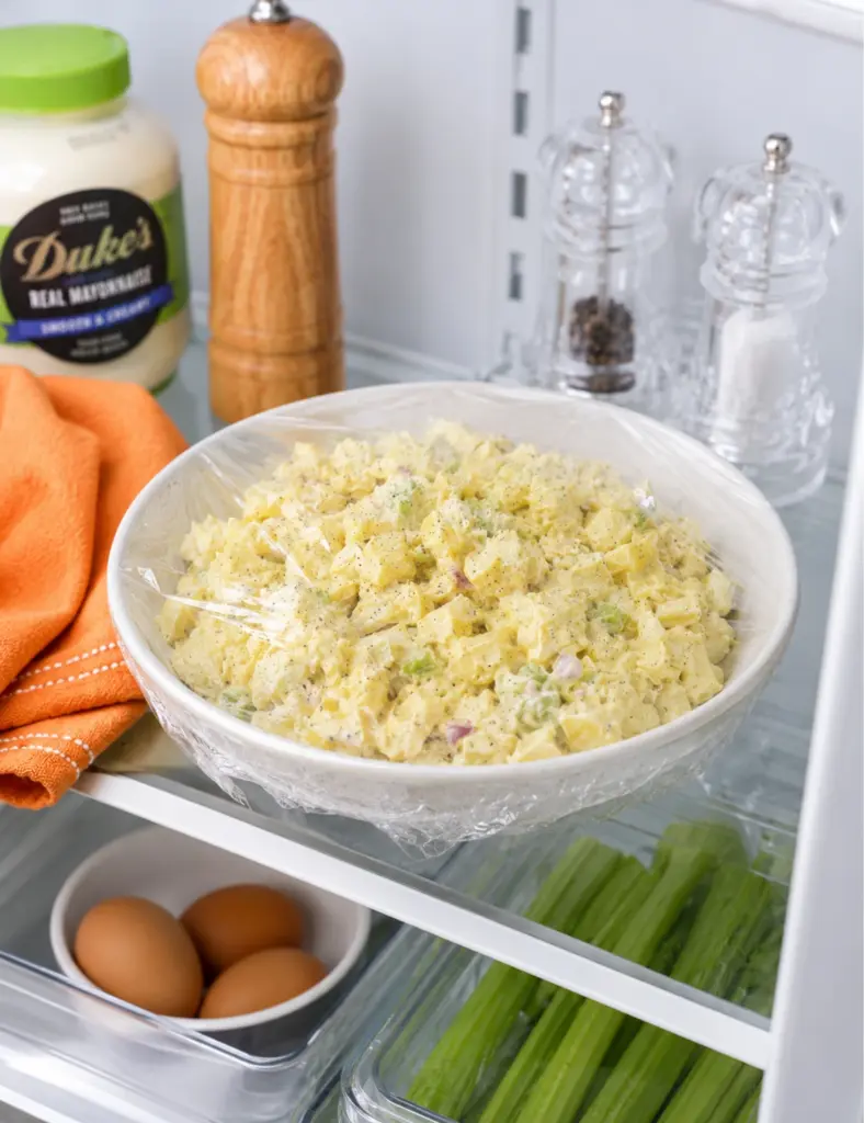 Classic potato salad covered with plastic wrap and chilling in the refrigerator, with creamy potatoes, celery, red onion, eggs, mayonnaise, and crystal shakers nearby.