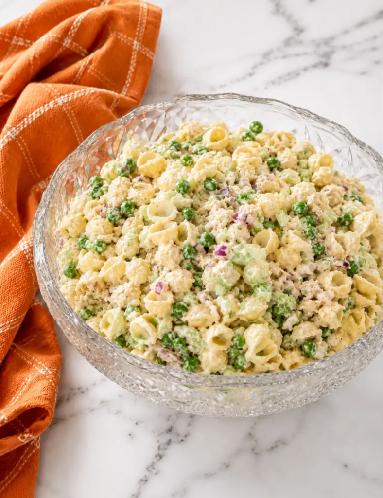 Finished tuna pasta salad with shell pasta, tuna, peas, celery, red onion, and creamy dressing in a crystal bowl on a marble countertop.
