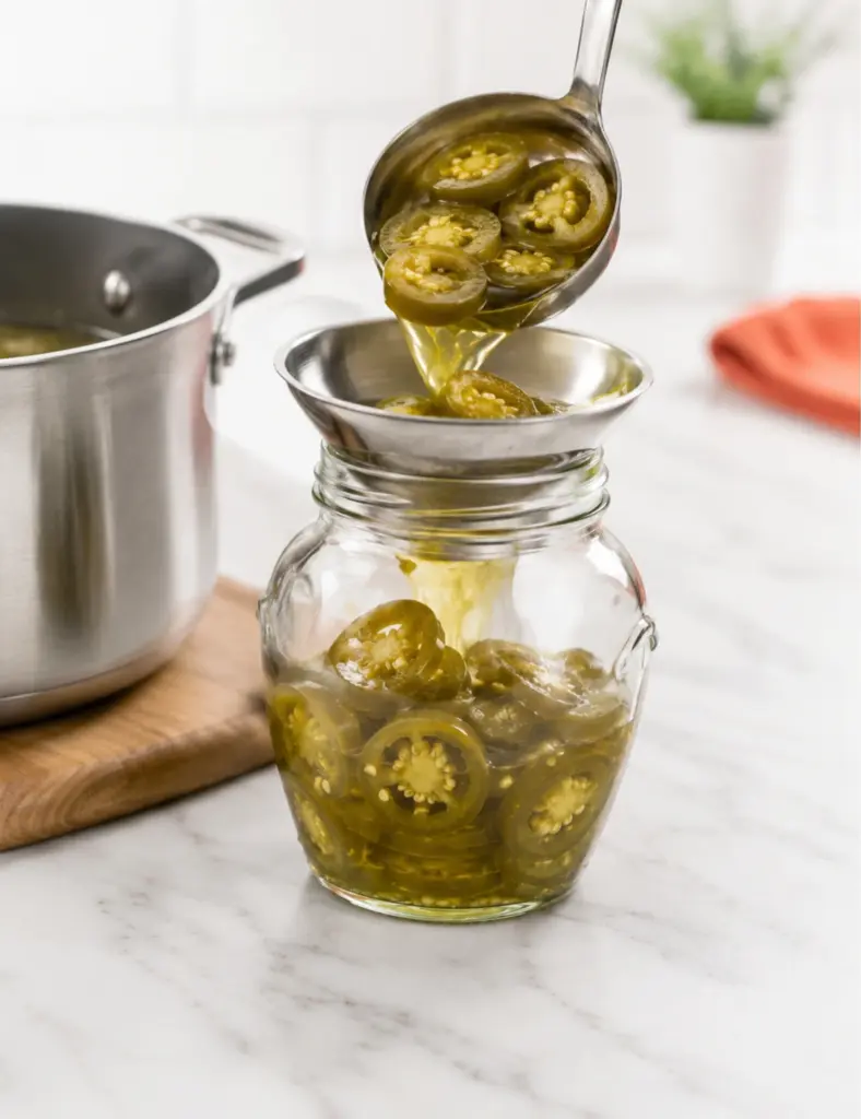 Hot Quick Pickled Jalapeños being ladled through a funnel into a glass jar with tangy brine on a white marble kitchen countertop.