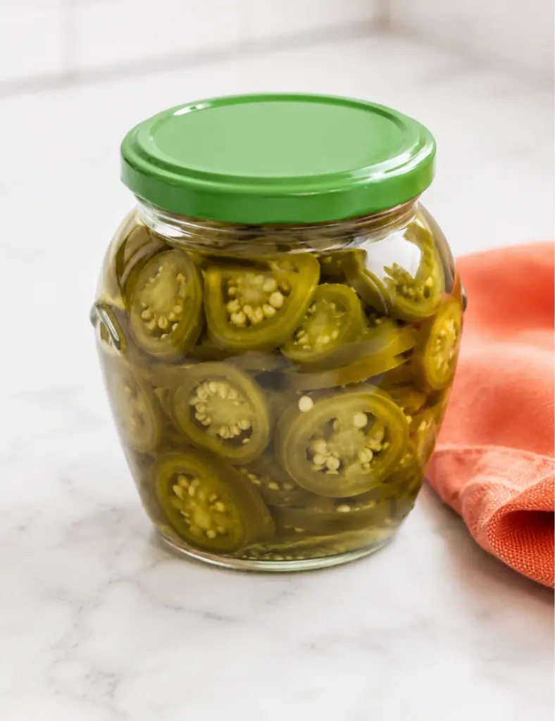 Jar of Quick Pickled Jalapeños with sliced green peppers in brine on a clean marble kitchen countertop with an orange towel nearby.