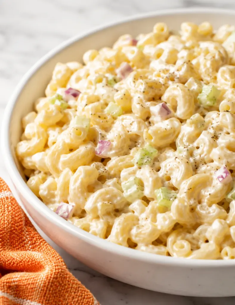 Close-up of creamy macaroni salad with elbow pasta, celery, red onion, egg, and black pepper in a white bowl on a marble countertop.