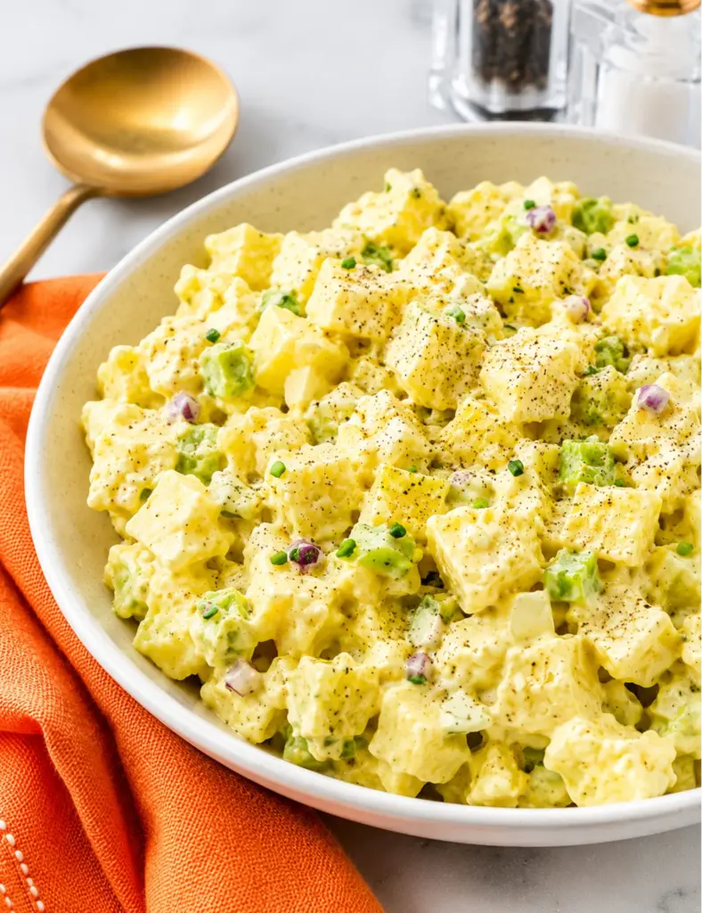 Close-up of Classic Potato Salad in a white bowl with creamy mustard dressing, Yukon Gold potatoes, celery, red onion, black pepper, and an orange kitchen towel.