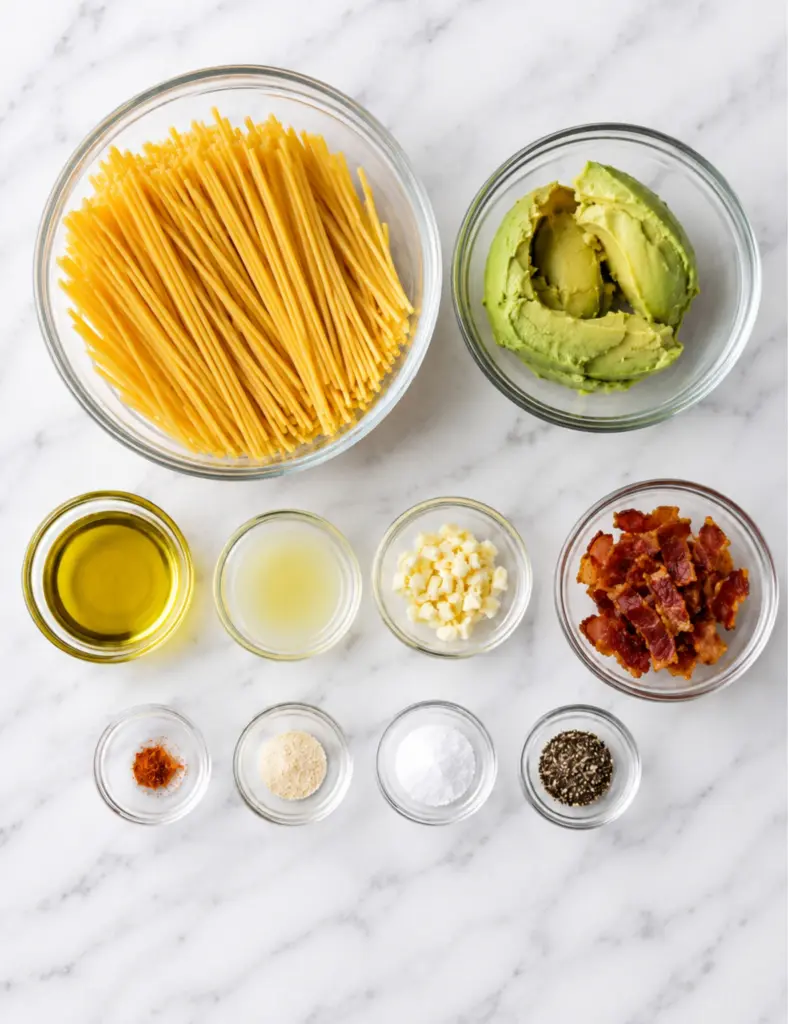 Avocado pasta ingredients in glass bowls with spaghetti, ripe avocado, olive oil, lemon juice, garlic, bacon, and seasonings on a white marble countertop