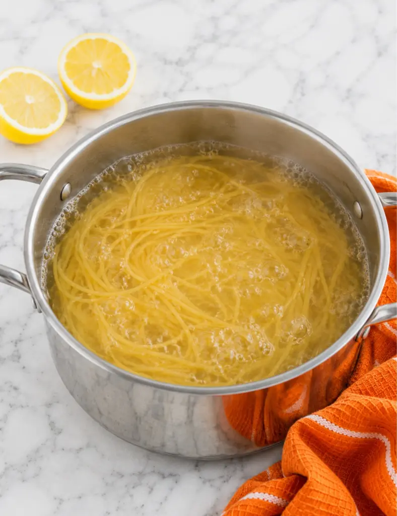 Spaghetti cooking in a stainless steel pot for avocado pasta on a white marble countertop with lemon halves and an orange kitchen towel