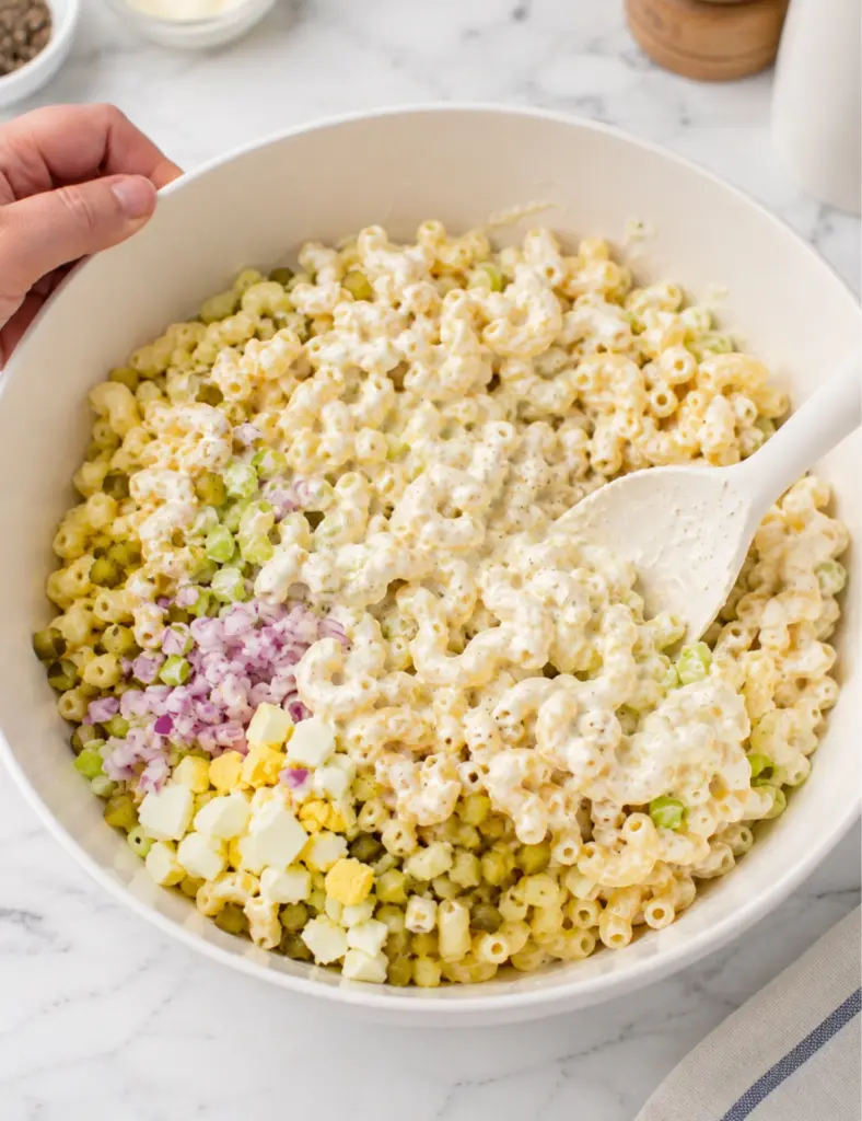 Macaroni salad being mixed in a large white bowl with elbow pasta, chopped eggs, pickles, celery, red onion, and creamy dressing.