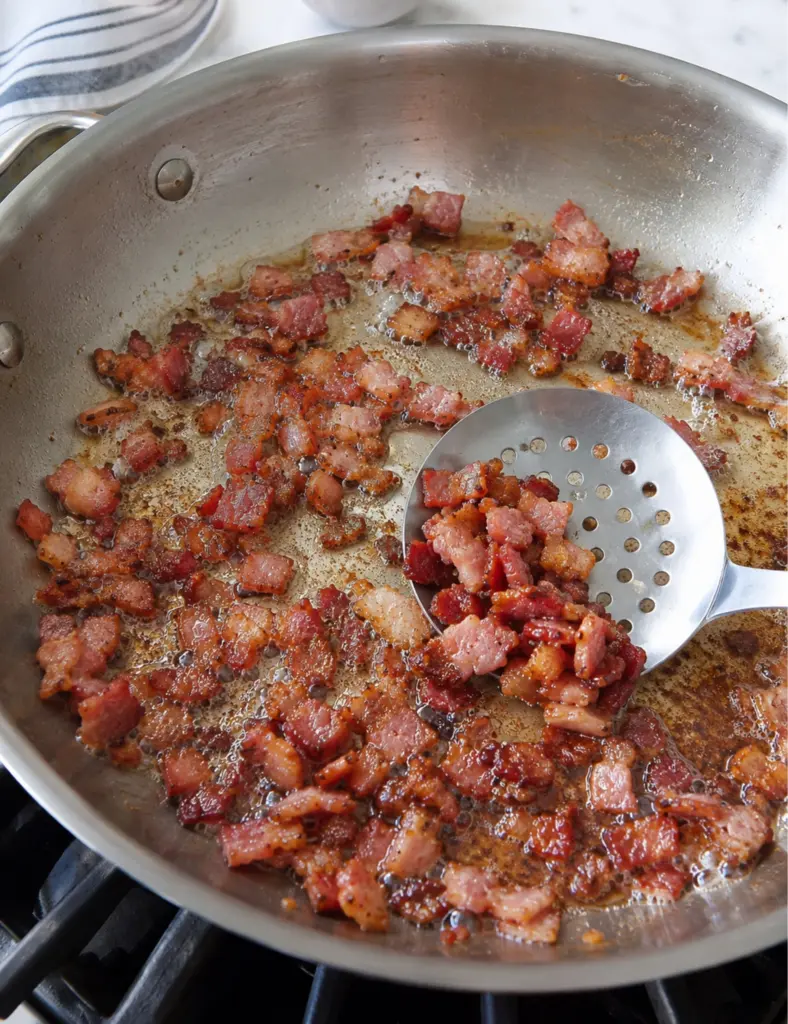 Crispy diced bacon cooking in a stainless steel pan with rendered fat for a homemade baked beans recipe.